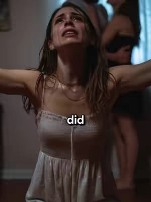 A young woman is depicted in a state of distress, with tears streaming down her face as she looks upward. She is wearing a light-colored, possibly white, slip dress, suggestive of sleepwear or lingerie. The background behind her is somewhat blurred, with other blurred figures partially visible, suggesting a scene within a room. The lighting appears to be dim, which adds to the dramatic mood.