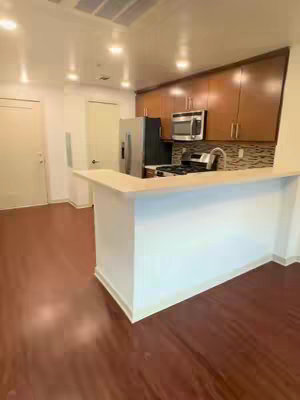 An interior shot of a modern kitchen with a breakfast bar and a partially visible stainless steel refrigerator. The kitchen area features dark wooden cabinets above and a mosaic tile backsplash by the cooking area. The floor is covered in wood-look laminate. The scene is brightly lit, suggesting a modern and well-maintained living space.