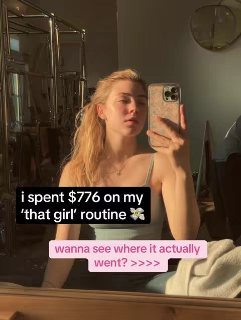 A young woman taking a selfie in a room. She is holding her phone up in front of her face, partially obscuring her features. She has long blond hair and is wearing a light-colored tank top. Behind her is a dark metal rack and a wall with sunlight streaming through. Text overlays are on the image, including a question about where her money went.