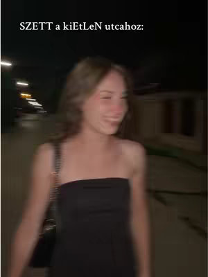 A night-time shot featuring a young woman walking along a pathway, brightly lit with overhead lights. She's wearing a black, strapless dress. The setting appears to be a street or walkway, with blurred details in the background suggesting movement and depth.