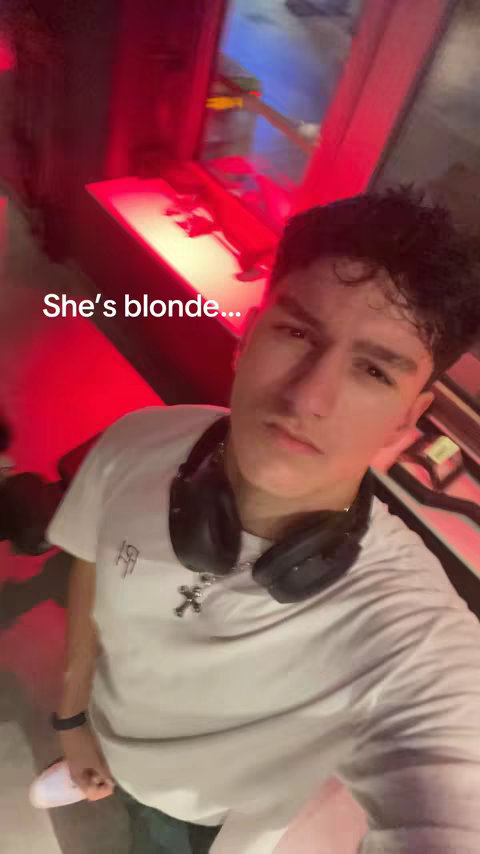 A selfie-style shot featuring a young man, centered and looking towards the camera with a neutral expression. He is wearing a white t-shirt and has dark hair. Black headphones are draped around his neck. Text overlayed on the image reads 'She's blonde...' The background is blurry with red and blue hues suggesting an indoor setting.