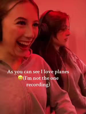 The image shows two young women, one in the foreground enthusiastically laughing with her head angled towards the camera, and another in the background with headphones on looking away. The scene is bathed in a red light.