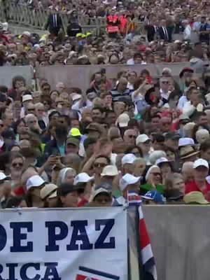 A crowded outdoor scene showing a sea of people. In the foreground, a banner with Spanish text is visible, and a partial flag can be seen in the bottom right corner. The crowd is tightly packed, and many people are holding up phones to capture the event.