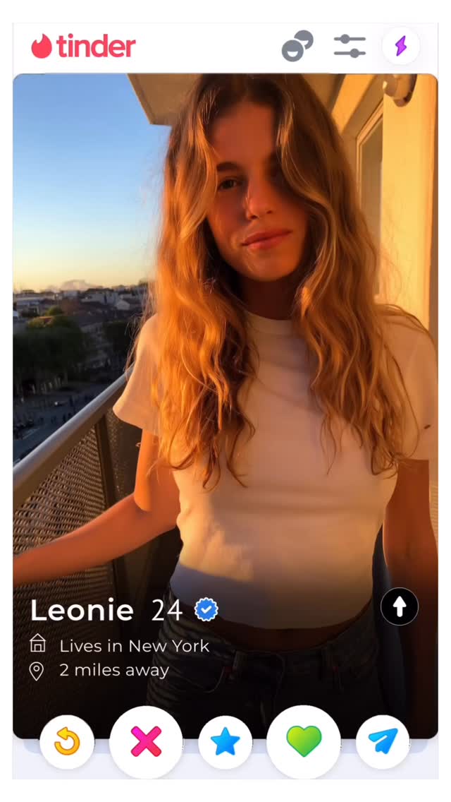 A vertical Tinder profile screenshot featuring a woman with long, wavy blonde hair. She stands on a balcony, the city and sunset visible behind her. The Tinder interface shows her name and age, along with details about her location and interests.