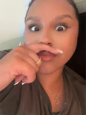A close-up shot of a person making a funny face. The person has dark skin, wide eyes, and a finger pressed against their upper lip, creating a fake mustache. They are wearing a top and a necklace.