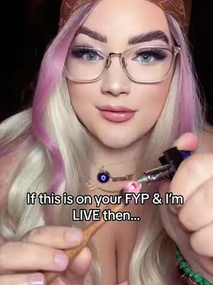 A close-up shot of a woman with fair skin, pink and blonde hair, and glasses holding a lighter and a small object. The woman is smiling slightly, with a brown scarf on her head. Text in white is overlaid on the image.