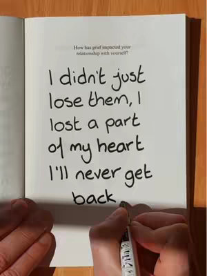 The image shows an open notebook with handwriting in black ink. The text reads, "I didn't just lose them, I lost a part of my heart I'll never get back." The hand of a person is visible, holding a pen and writing more text on the page. The notebook is open on a wooden surface.