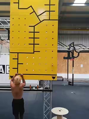 A man is seen from the back, shirtless, hanging from a yellow climbing wall with cut-out shapes. The wall dominates the frame, with the man's arms and upper back visible. The setting appears to be a gym or indoor training facility.