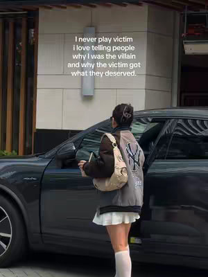 A person in a grey and brown jacket, white skirt, and white socks stands next to a dark grey car with a building in the background. Text is overlaid on the top left with white font.