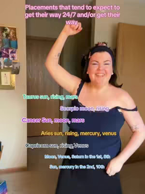 A woman is standing in a room with her arm raised, smiling. She's wearing a dark blue spaghetti-strap top. Text overlays list astrological placements in different colors.