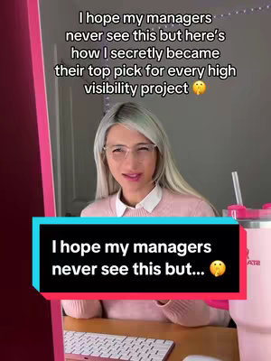 A woman wearing glasses, a light pink sweater, and a white collared shirt beneath is seated at a desk. The background includes a light-colored wall, hints of pink framing on the side, and a gray wall. Below that, a black rectangle with text overlaid. The woman has blonde hair and appears to be smiling slightly as she sits in front of the monitor, with a pink cup at the side.