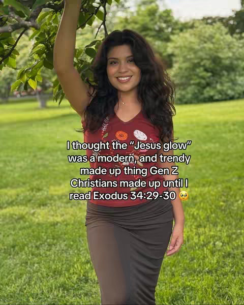 A young woman with long, dark hair and a smile is shown outdoors. She's wearing a red top and a dark skirt, with one arm raised up towards a tree branch. The background is a green grassy area, with trees and a bright sky suggesting a sunny day. Text is overlaid onto the image.