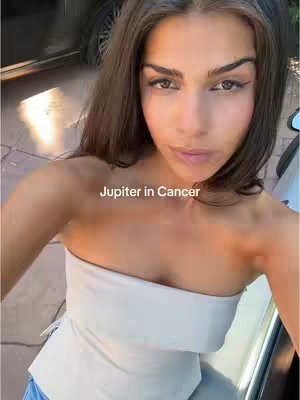 A selfie-style shot taken from the chest up of a young woman with long brown hair. She is wearing a white strapless top and has makeup on, including eyeliner. The background shows what looks like the inside and outside of a car. Text overlayed onto the image says "Jupiter in Cancer".