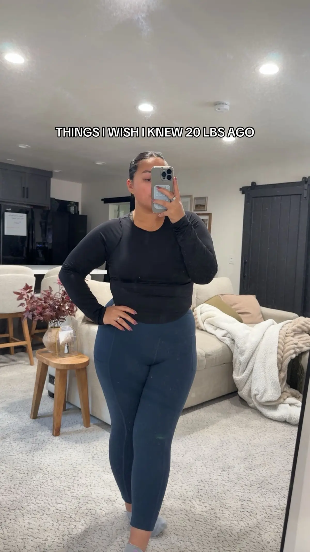 A woman is taking a selfie in a well-lit room. She is standing in front of a large mirror, holding up her phone to capture the image. She is wearing a black long-sleeved shirt and blue leggings. The background shows a living room with a couch.