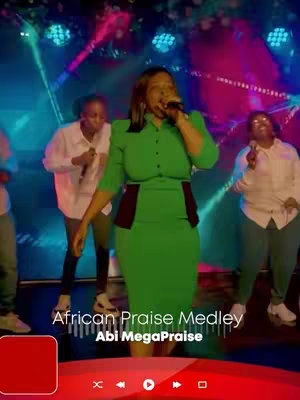 A live performance shot featuring a female singer, center stage, wearing a green dress. She holds a microphone and is actively singing. Behind her are background singers and a stage setup with colorful lights. Below the image is text identifying the song and artist.