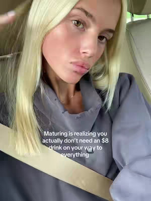 A selfie-style shot featuring a young woman inside a car. The woman, with light skin and blonde hair, looks towards the camera. She is wearing a grey hoodie and a seatbelt is visible across her torso. Text overlayed on the image says 'Maturing is realizing you actually don't need an $8 drink on your way to everything'. The composition is framed from the chest up, concentrating on her face and upper body, with the car's interior serving as the environment.