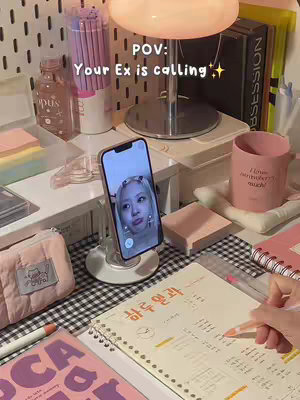 The cover image depicts a well-lit, cozy workspace. A smartphone in a holder displays a video call, with a woman's face visible on the screen. Surrounding the phone are desk essentials: a notebook with Korean writing, pink sticky notes, pens, a pink mug with text, and various decorative items.