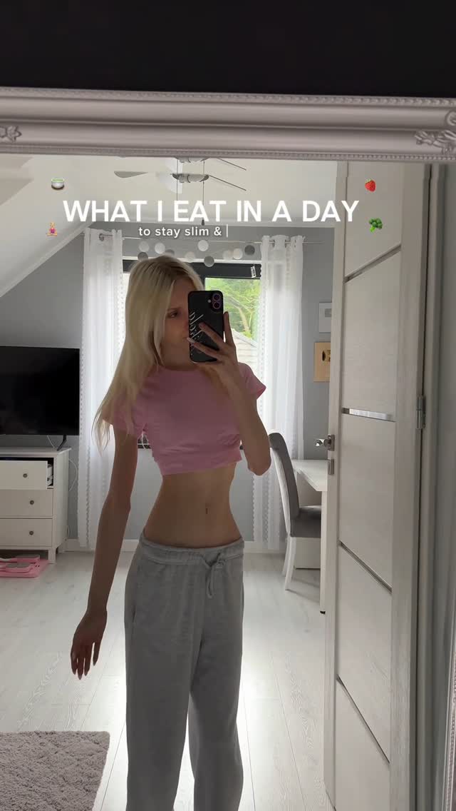 A young woman is taking a mirror selfie, showing her in a casual outfit in her bedroom. The image features text overlays and fruit emojis.