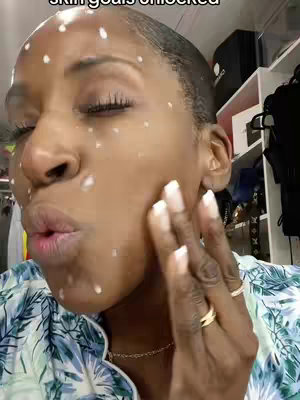 A close-up shot of a woman's face from the chest up, with skincare product dots on her face. She has her eyes closed and is touching her cheek with her fingers. The background includes a shelf with items and a closet.