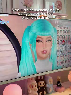The image displays the front of a computer monitor. On the screen, a character from The Sims 4 is visible, with teal pigtails and heavy makeup. Text overlaying the screen reads "Sims 4 girlies spending hours doing everything but playing the Sims." Below the monitor, there are small figurines and decorative items on a shelf.