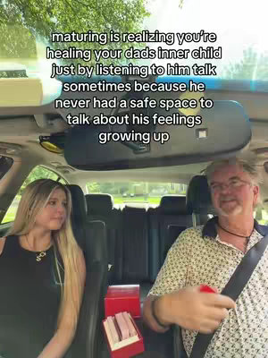 Two people sitting in the front seats of a car are filmed from inside the vehicle. The person on the left is a young woman with long blonde hair, wearing a black top with a necklace, looking slightly towards the other person. The person on the right is an older man wearing a patterned shirt, a dark collar, and glasses. Both are sitting in the front of a car. A text overlay is at the top.