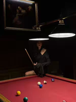 A woman in a dark room is poised with a pool cue over a red pool table, balls scattered across it. Overhead, dim lights cast shadows. A framed painting hangs on the wall. The image has a moody and dramatic feel.