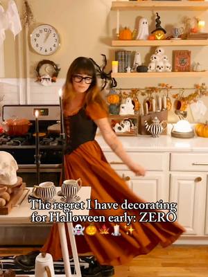 A woman in a Halloween-themed kitchen, wearing an orange dress and glasses, dances while holding a lighted candle.