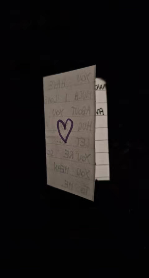 A close-up shot of a folded piece of paper against a black background. The paper has handwriting visible, with words and a purple heart drawn on it. The paper is positioned at an angle, partially open, with the edge slightly visible.