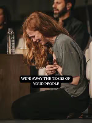 The image features a woman with long, red hair, holding a microphone and crying. She is kneeling, with her hands clasped near her chest. The background is slightly blurred, suggesting a stage or event setting. A man in a dark suit is visible in the background. A text overlay reads "WIPE AWAY THE TEARS OF YOUR PEOPLE". The lighting appears to be warm, creating a somber mood.