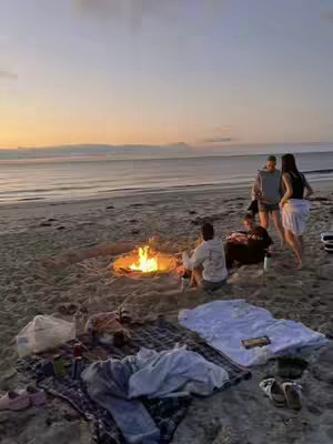 A photograph captures a beach scene at dusk. In the foreground, a sandy beach is scattered with blankets, a pile of clothes, and shoes. In the middle ground, a bonfire is lit, with a group of people sitting around it. A calm ocean stretches into the background, reflecting the colors of the sunset, with shades of orange and pink.