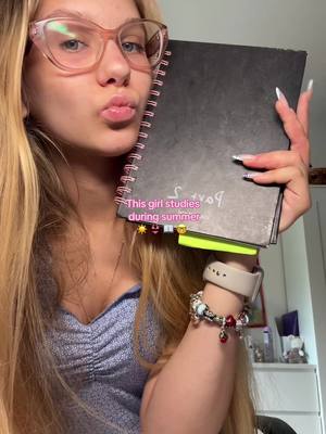 A young woman with long blonde hair, wearing glasses and a light blue top poses, holding a spiral notebook and making a kissing face. Text overlayed on the notebook says "This girl studies during summer".