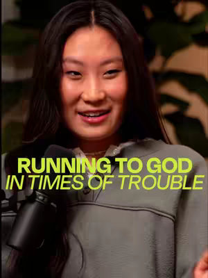 A woman with long, dark hair is centered in the frame, smiling and facing the camera. The background is softly blurred, with hints of green foliage visible on the sides. The woman wears a gray sweatshirt. Large, neon-green text overlays her, and another microphone is visible.