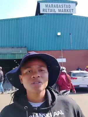 A woman in a black hoodie and a blue hat stands in front of a building that says 'MARABASTAD RETAIL MARKET' on it. There are other people in the background.