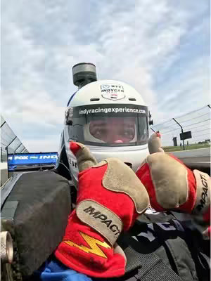 A first-person view from inside an IndyCar race car. The driver, wearing a white helmet with sponsor logos, red and tan gloves, and a black racing suit, gives a thumbs-up with both hands. The background shows the racetrack with blue sky and scattered clouds.