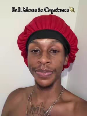 A selfie-style image featuring a young Black man against a white background. He's wearing a red bonnet and a black durag underneath. He's looking directly at the camera with his tongue slightly sticking out, and has a subtle cross tattoo visible. The text 'Full Moon in Capricorn' is overlaid at the top.