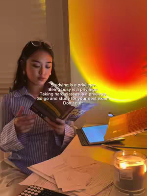 A young woman with dark hair wearing glasses on top of her head is sitting at a desk in a room lit by a sunset lamp. She is holding a book in her hands and looking down at it. There are papers strewn across the desk, along with a tablet, laptop and a candle. Text overlays are written on the top and side of the image, offering both encouragement and motivation.