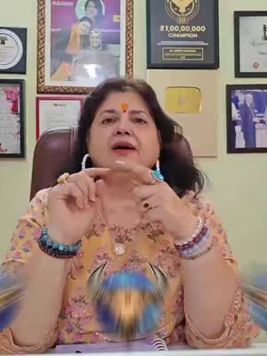 A woman is seated, facing the camera. She gestures with her hands and has a red bindi on her forehead. The background includes framed pictures and a YouTube Play Button award.