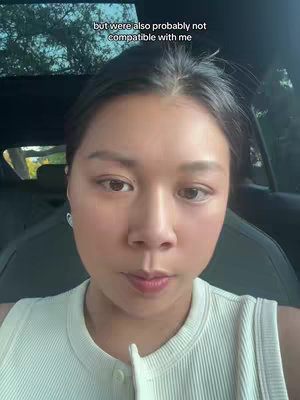 A selfie taken from inside a car. The focus is on a woman's face, her eyes looking off to the side. Behind her, the interior of the car is visible, and through the window blurred trees are seen. The lighting is somewhat soft and natural. The woman wears a simple, light-colored top.