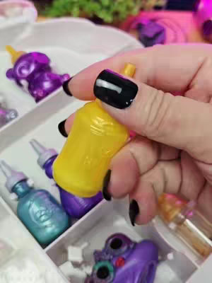 A close-up shot of a hand holding a yellow plastic container, possibly a container for slime. The hand has very dark, glossy black nail polish. Other colorful containers and slime toys are visible in the background, set in a white segmented tray. The color palette includes yellow and other vibrant colors, with a shallow depth of field focusing on the hand and container.