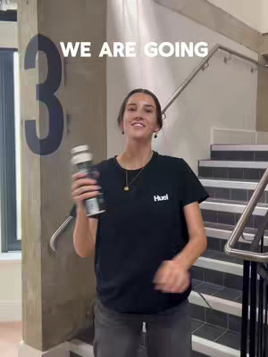 A woman stands in a well-lit stairwell, holding a bottle while smiling at the camera. Behind her is a large number 3, and text overlays read 'WE ARE GOING'. The background includes a handrail and stairs.