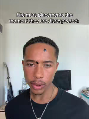 A medium shot features a young Black man as the primary focus. He has short, neatly styled dark hair. Two small, dark star designs are placed on his forehead, one centered and the other slightly above and to the side. He is wearing a black t-shirt and a silver chain around his neck. The background is simple, with a white wall, a partial view of a guitar, and the corner of a television screen. Above the man's head is text.