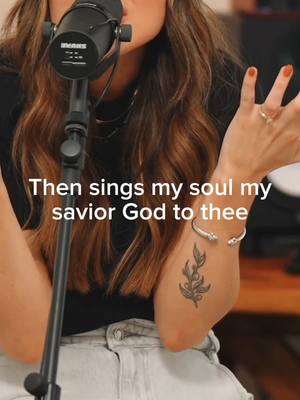 The cover image features a medium close-up of a person singing into a microphone. The individual is centered in the frame, with the microphone obscuring part of their face. They have long, wavy brown hair. The person is wearing a black top and light-colored jeans. Visible are a silver bracelet, several rings and a tattoo on their left arm. The background is blurred, but appears to be a room setting with a wooden surface and some visible objects.