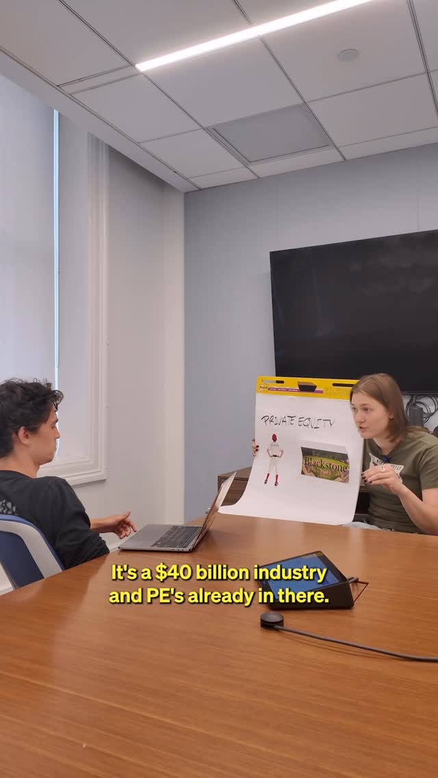 The image shows two people in a meeting room. One person, a young woman, is holding up a whiteboard with the words 'PRIVATE EQUITY' written on it, along with a stick figure, a diagram and the Blackstone logo. The other person, a young man, is seated at a table with an open laptop is looking at the whiteboard and woman.  There is a table in the foreground with a black device on it.