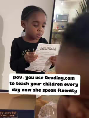 A young girl is holding a paper with the word "MANGALA" written on it. She is looking at the paper and appearing to read it. The shot is from a slightly low angle, with the girl filling the majority of the frame. The background is blurred, with glimpses of a room visible and what looks like a person behind her. There is a text overlay at the bottom that reads "pov: you use Reading.com to teach your children every day now she speak fluently".