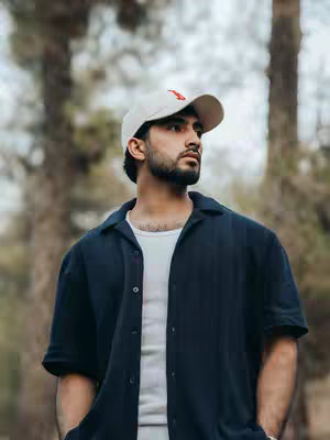 A man in a white cap with a red logo is looking to the side. He is wearing a dark blue button-down shirt over a white t-shirt. The background is blurred, featuring trees and foliage.