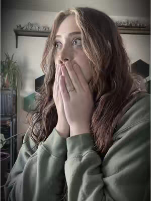 A woman with long, wavy brown hair with blonde highlights is in a room, with a surprised expression, covering her mouth with both hands as if in shock. She’s wearing a casual green hoodie. The background includes out-of-focus decor with neutral colors.