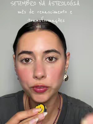A selfie-style shot features a woman from the chest up, looking directly at the camera. The background is a neutral wall. Overlaid atop the image is white text in a handwritten-style font. The woman has fair skin, dark hair, and subtle makeup including blush. She wears dark t-shirt and necklace. A yellow smiley face is in her hand. She is also wearing earrings.