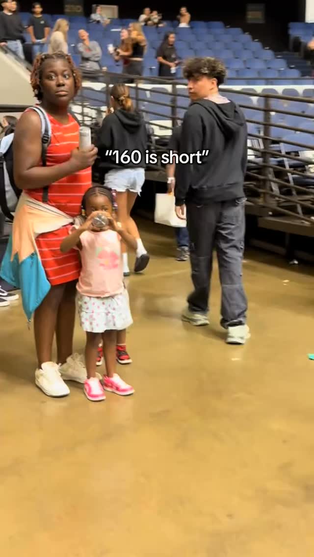 The cover image captures a scene at what appears to be an event or venue, with a crowd in the background and foreground figures. A  person of color in a red and white striped dress stands on the left, with a young child holding a clear bottle partially obstructing their face. Another figure, a young man wearing a black hooded sweatshirt, is visible, and text overlayed: "160 is Short" is displayed in white text. The scene is lit by bright lighting, and the setting seems casual and energetic.