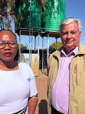 Two people are standing in front of two large green water tanks. A diverse pair is standing side-by-side, with a person wearing glasses and a white top on the left, and a person in a beige jacket on the right.