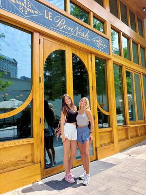 Two young women stand in front of a wooden-framed building with large glass windows and arched doorways, the sign overhead reads 'LE Bon Nosh'. The women are smiling and wearing casual summer outfits, one in a black top and white shorts and the other in a gray tank top and blue shorts.